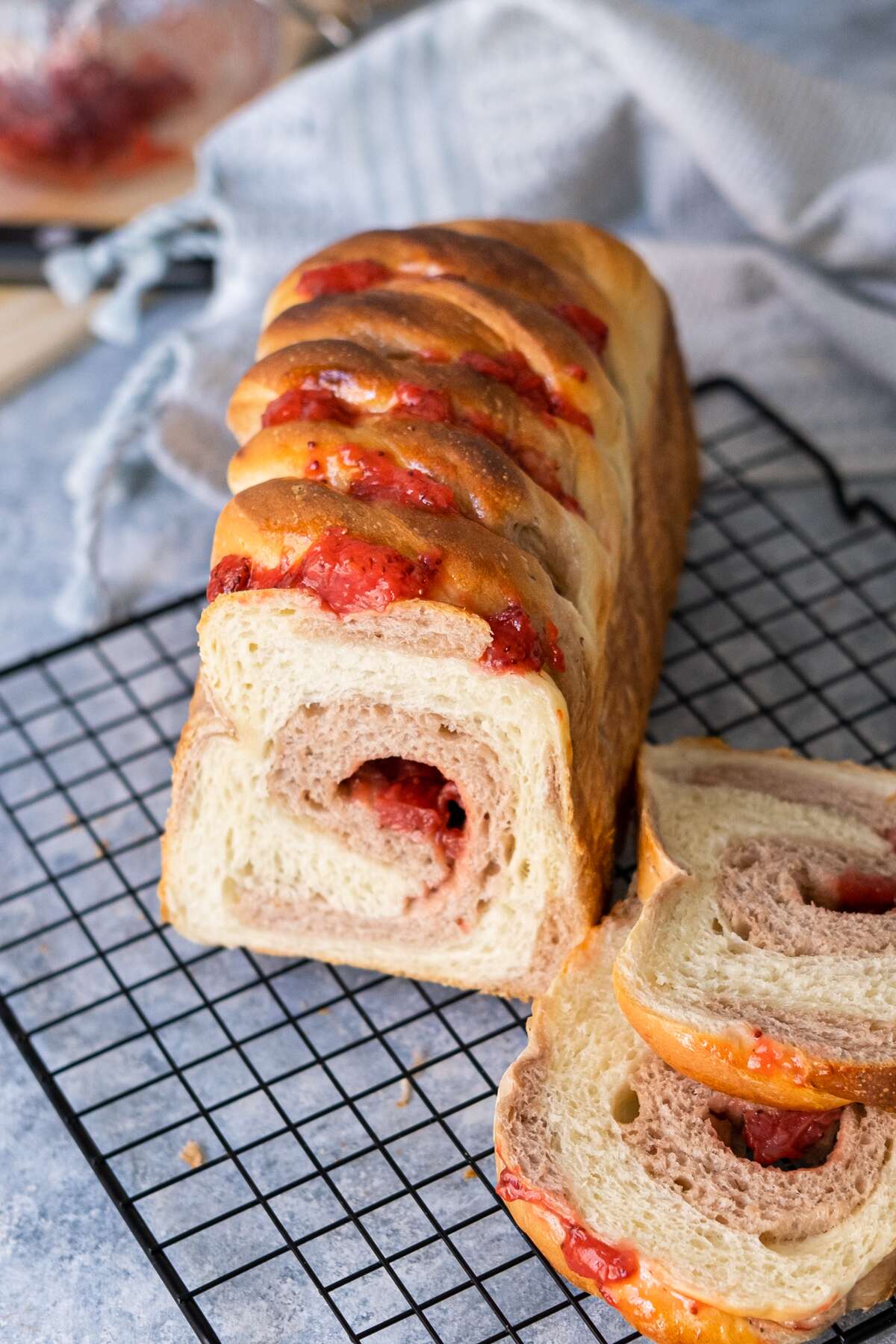 A loaf of strawberry Brioche with two slices on the wire rack with blue kitchen towel aside.
